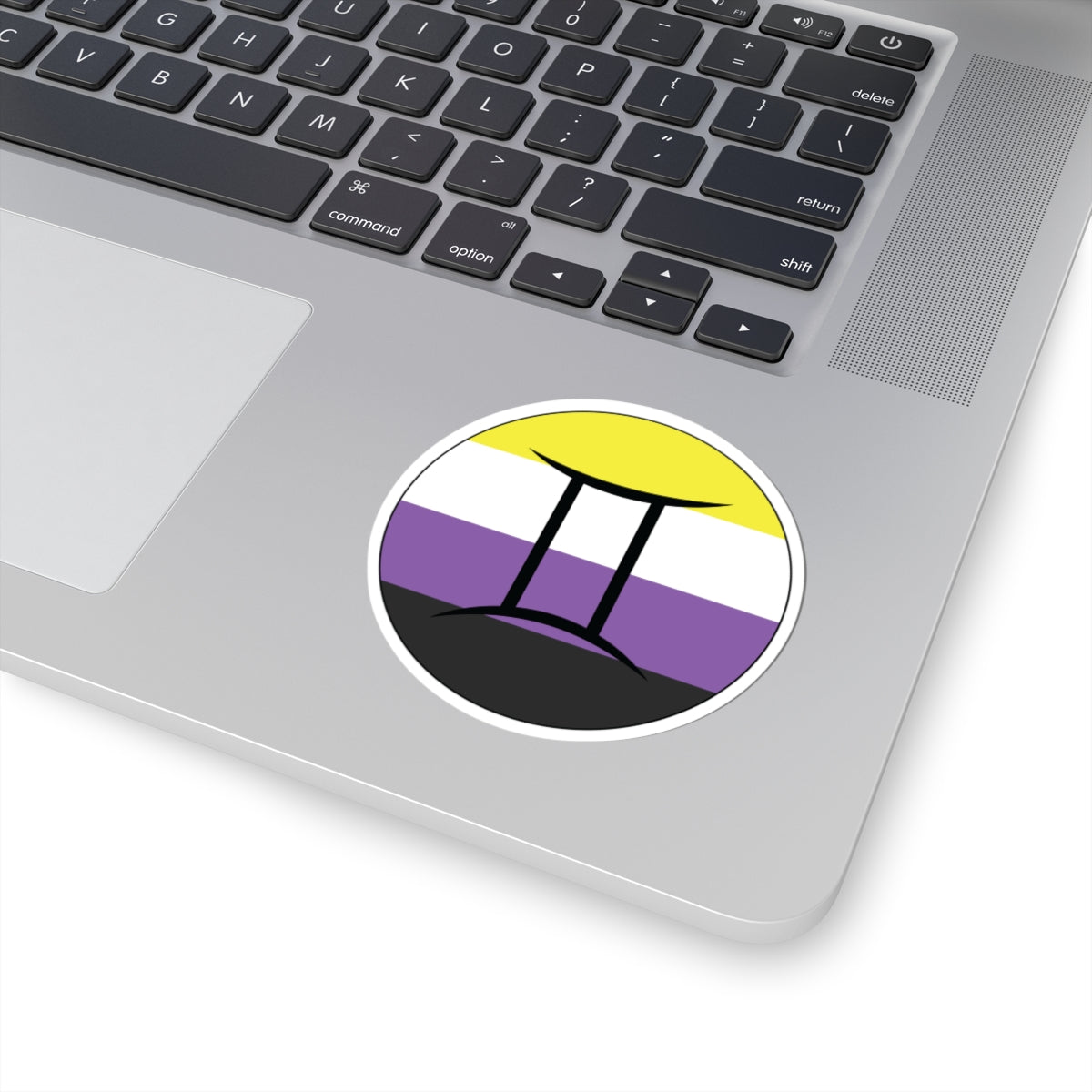 Close-up of the bottom right corner of a silver laptop featuring a 3 inch by 3 inch circular zodiac sticker with a centered Gemini glyph on a Nonbinary pride flag style background applied to the surface.