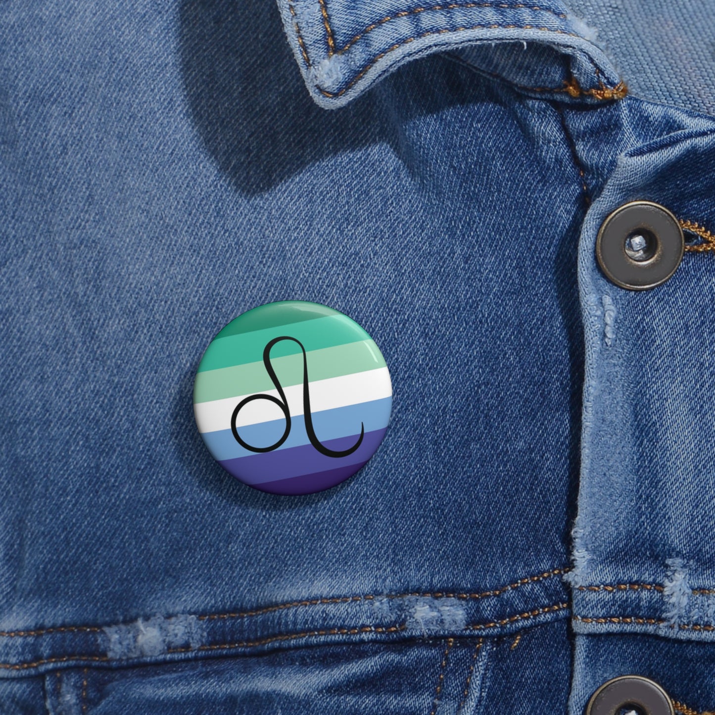 Close-up of a white circular pride button featuring the Leo zodiac glyph centered over a gay men (MLM) pride flag background in greens/teals, white, and blues, attached to a blue denim jacket at the right chest with the button clearly visible.