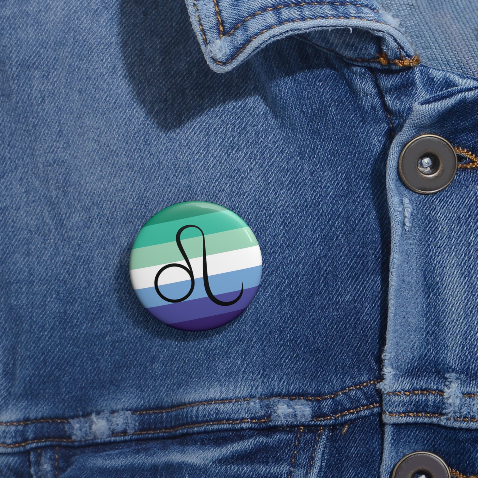 Close-up of a white circular pride button featuring the Leo zodiac glyph centered over a gay men (MLM) pride flag background in greens/teals, white, and blues, attached to a blue denim jacket at the right chest with the button clearly visible.