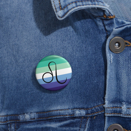 Close-up of a white circular pride button featuring the Leo zodiac glyph centered over a gay men (MLM) pride flag background in greens/teals, white, and blues, attached to a blue denim jacket at the right chest with the button clearly visible.