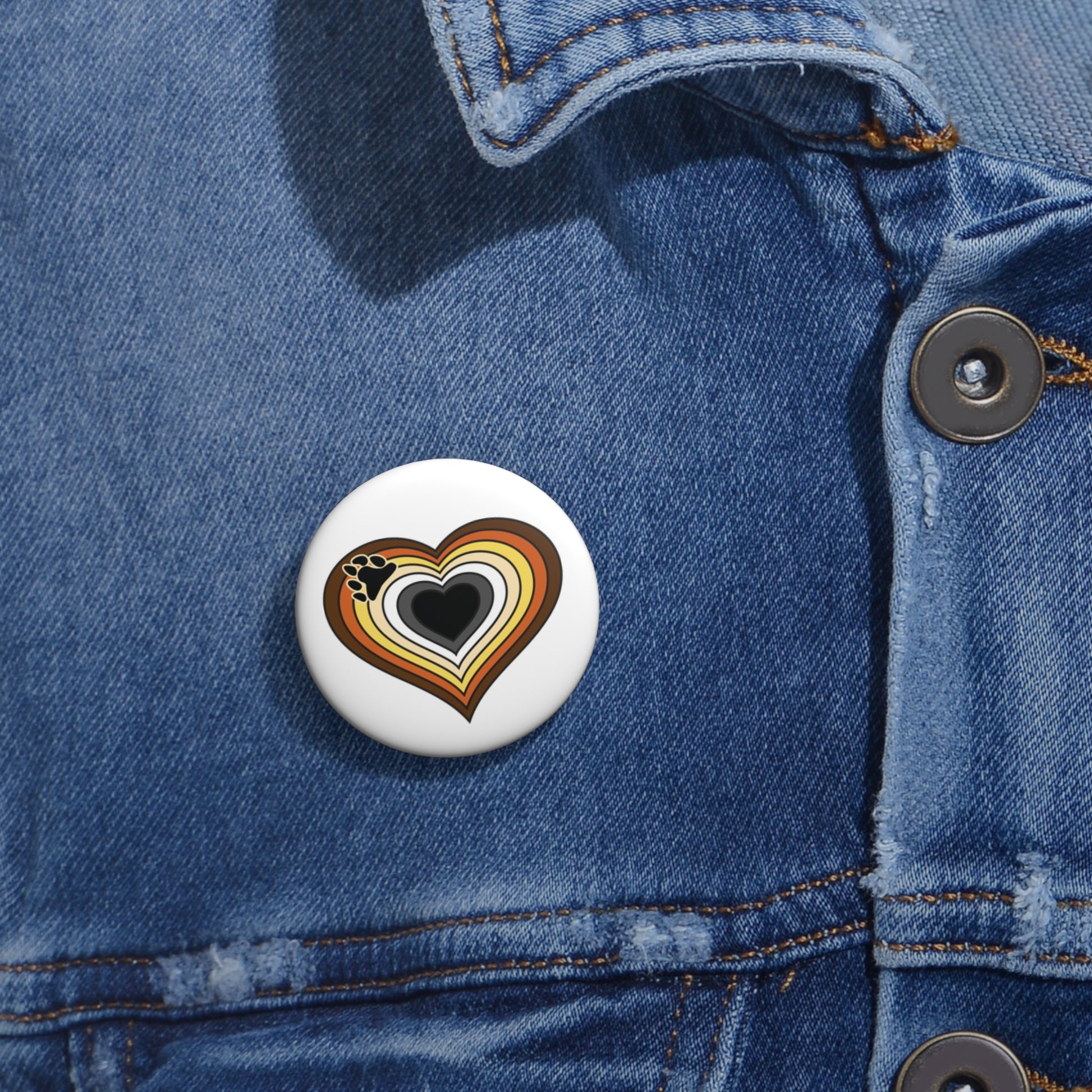 Close-up of a white circular pride button featuring a nested heart design inspired by the Bear pride flag in browns, cream, white, grey and black, attached to a blue denim jacket at the right chest with the button clearly visible.