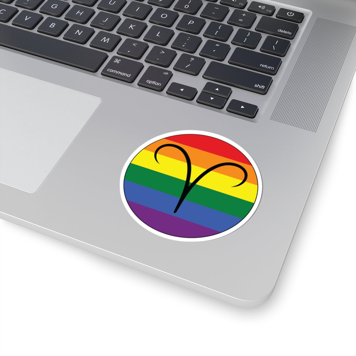 Close-up of the bottom right corner of a silver laptop featuring a 3 inch by 3 inch circular zodiac sticker with a centered Aires glyph on a Rainbow pride flag style background applied to the surface.