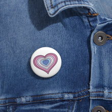 Close-up of a white circular pride button featuring a nested heart design inspired by the Bigender pride flag in pinks, lavender, white and blues, attached to a blue denim jacket at the right chest with the button clearly visible.