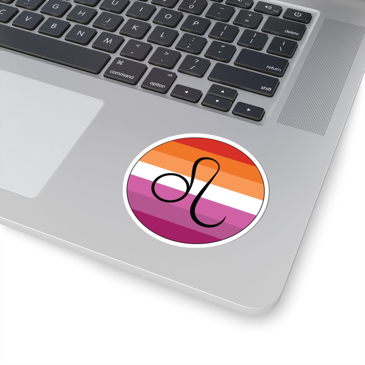Close-up of the bottom right corner of a silver laptop featuring a 3 inch by 3 inch circular zodiac sticker with a centered Leo glyph on a Lesbian pride flag style background applied to the surface.