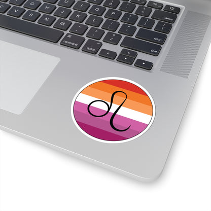 Close-up of the bottom right corner of a silver laptop featuring a 3 inch by 3 inch circular zodiac sticker with a centered Leo glyph on a Lesbian pride flag style background applied to the surface.