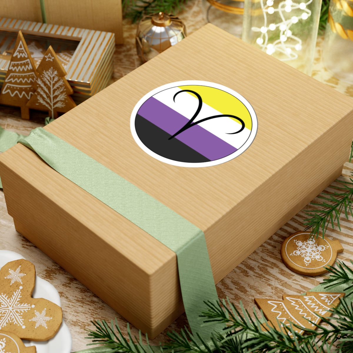Gift box with a green ribbon on a festive background with cookies and Christmas decorations. Aires Nonbinary pride sticker centered on the box.