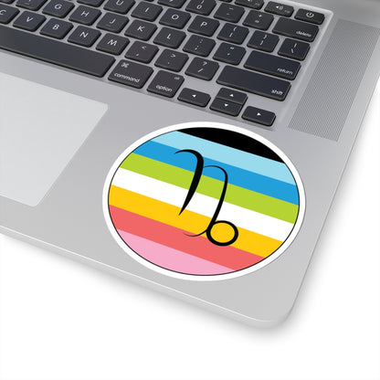 Close-up of the bottom right corner of a silver laptop featuring a 4 inch by 4 inch circular zodiac sticker with a centered Capricorn glyph on a Queer pride flag style background applied to the surface.