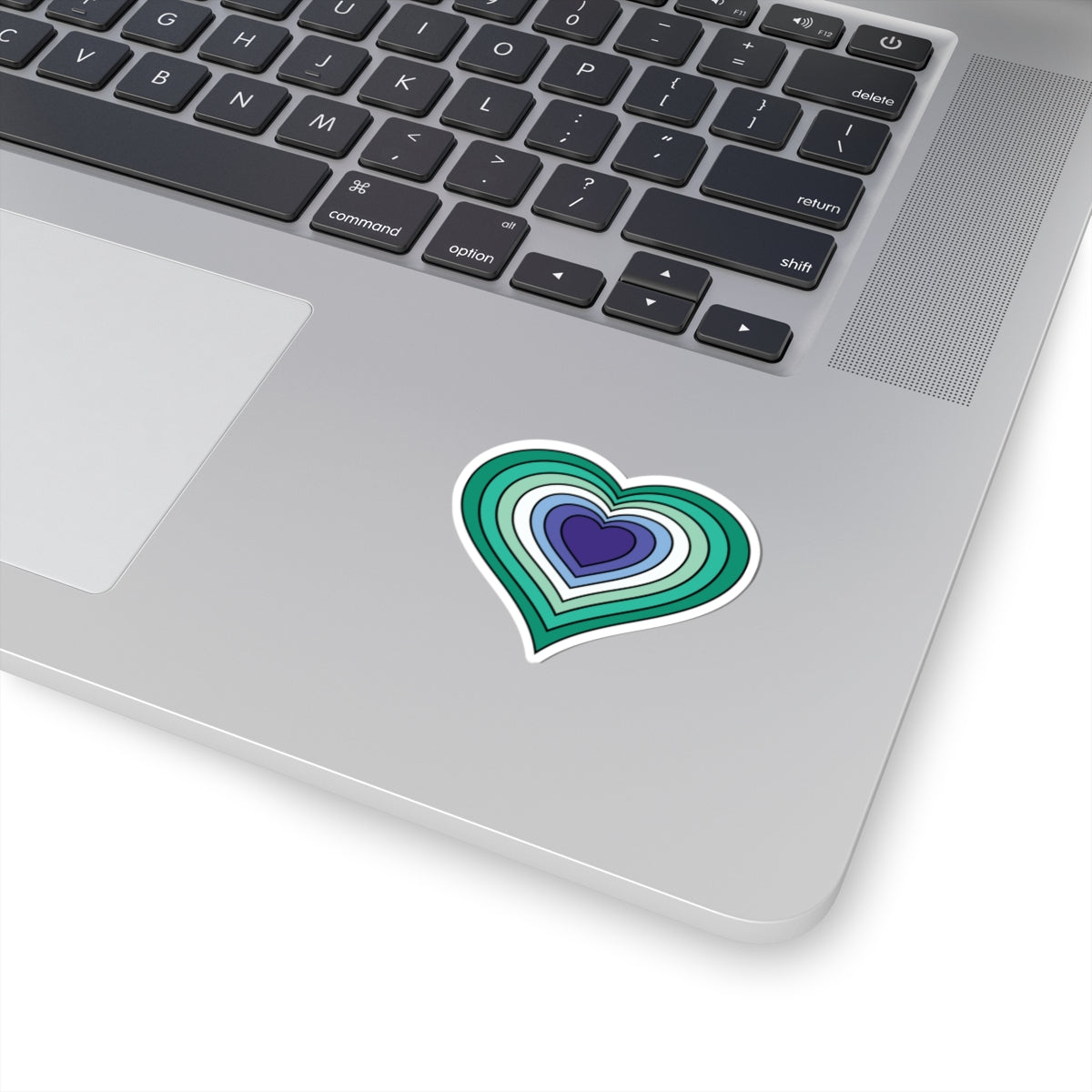 Close-up of the bottom right corner of a silver laptop featuring a 2 inch by 2 inch pride flag heart sticker in a Gay Men flag style applied to the surface.