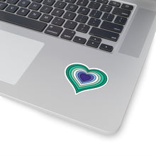 Close-up of the bottom right corner of a silver laptop featuring a 2 inch by 2 inch pride flag heart sticker in a Gay Men flag style applied to the surface.
