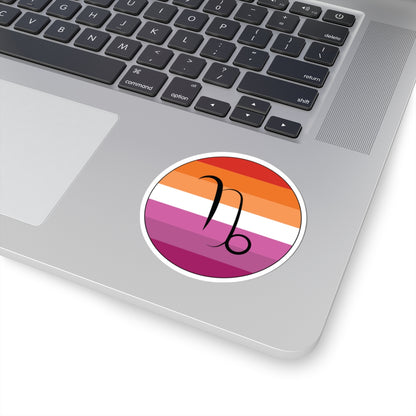 Close-up of the bottom right corner of a silver laptop featuring a 3 inch by 3 inch circular zodiac sticker with a centered Capricorn glyph on a Lesbian pride flag style background applied to the surface.
