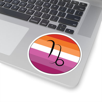 Close-up of the bottom right corner of a silver laptop featuring a 4 inch by 4 inch circular zodiac sticker with a centered Capricorn glyph on a Lesbian pride flag style background applied to the surface.