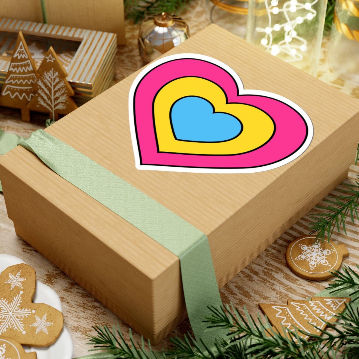Gift box with a green ribbon on a festive background with cookies and Christmas decorations. Heart-shaped Pansexual pride sticker centered on the box.