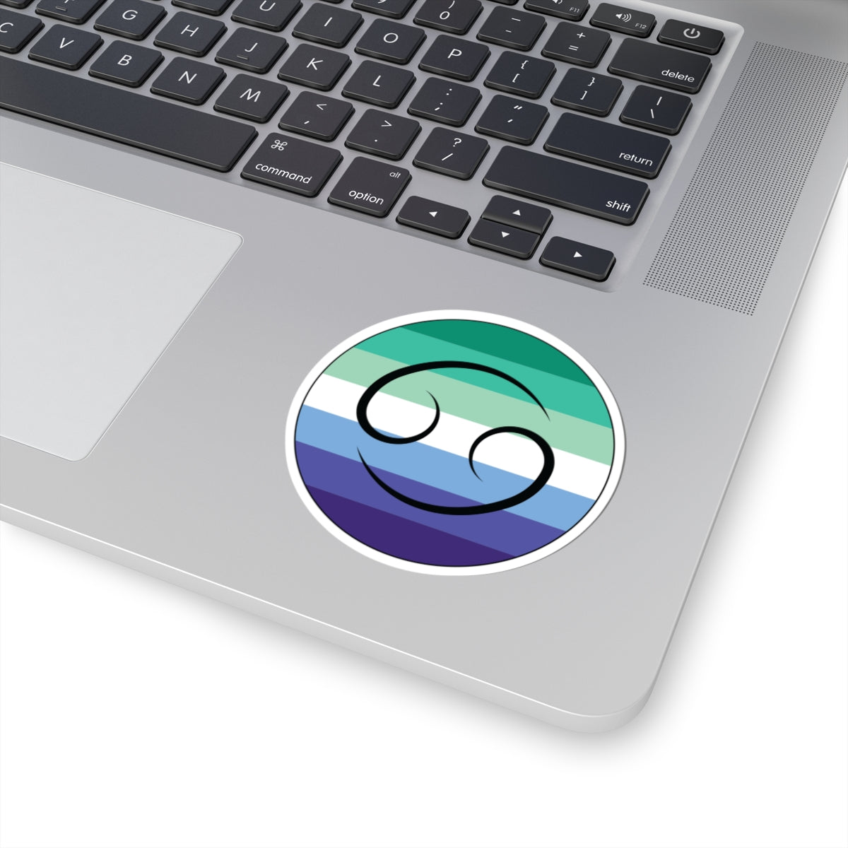 Close-up of the bottom right corner of a silver laptop featuring a 3 inch by 3 inch circular zodiac sticker with a centered Cancer glyph on a Gay Men pride flag style background applied to the surface.
