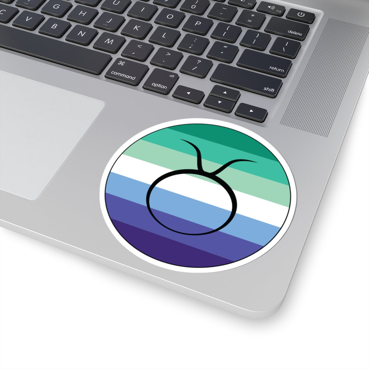 Close-up of the bottom right corner of a silver laptop featuring a 4 inch by 4 inch circular zodiac sticker with a centered Taurus glyph on a Gay Men pride flag style background applied to the surface.