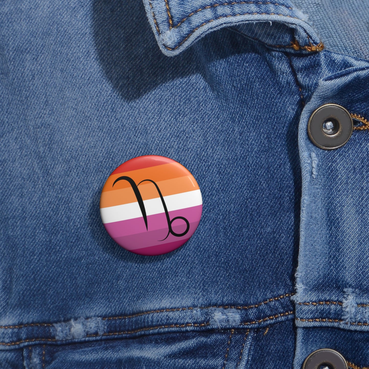 Close-up of a white circular pride button featuring the Capricorn zodiac glyph centered over a lesbian pride flag background in oranges, white, and pinks attached to a blue denim jacket at the right chest with the button clearly visible.