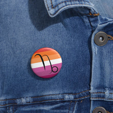 Close-up of a white circular pride button featuring the Capricorn zodiac glyph centered over a lesbian pride flag background in oranges, white, and pinks attached to a blue denim jacket at the right chest with the button clearly visible.