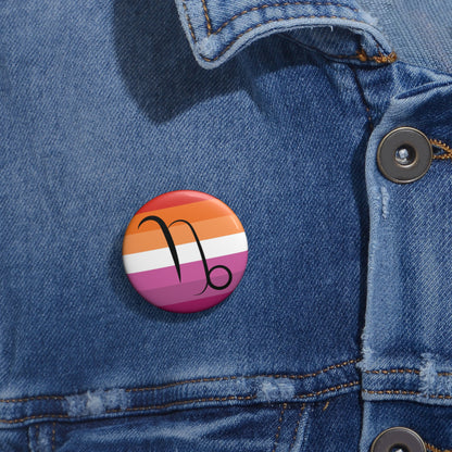 Close-up of a white circular pride button featuring the Capricorn zodiac glyph centered over a lesbian pride flag background in oranges, white, and pinks attached to a blue denim jacket at the right chest with the button clearly visible.