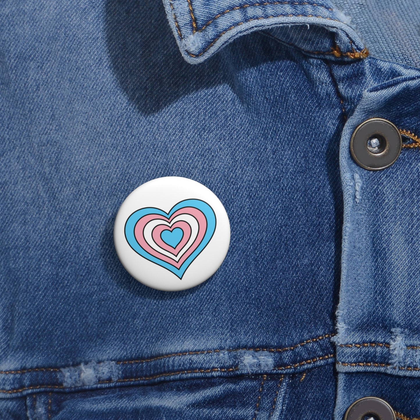 Close-up of a white circular pride button featuring a nested heart design inspired by the Transgender pride flag in blue, pink and white, attached to a blue denim jacket at the right chest with the button clearly visible.
