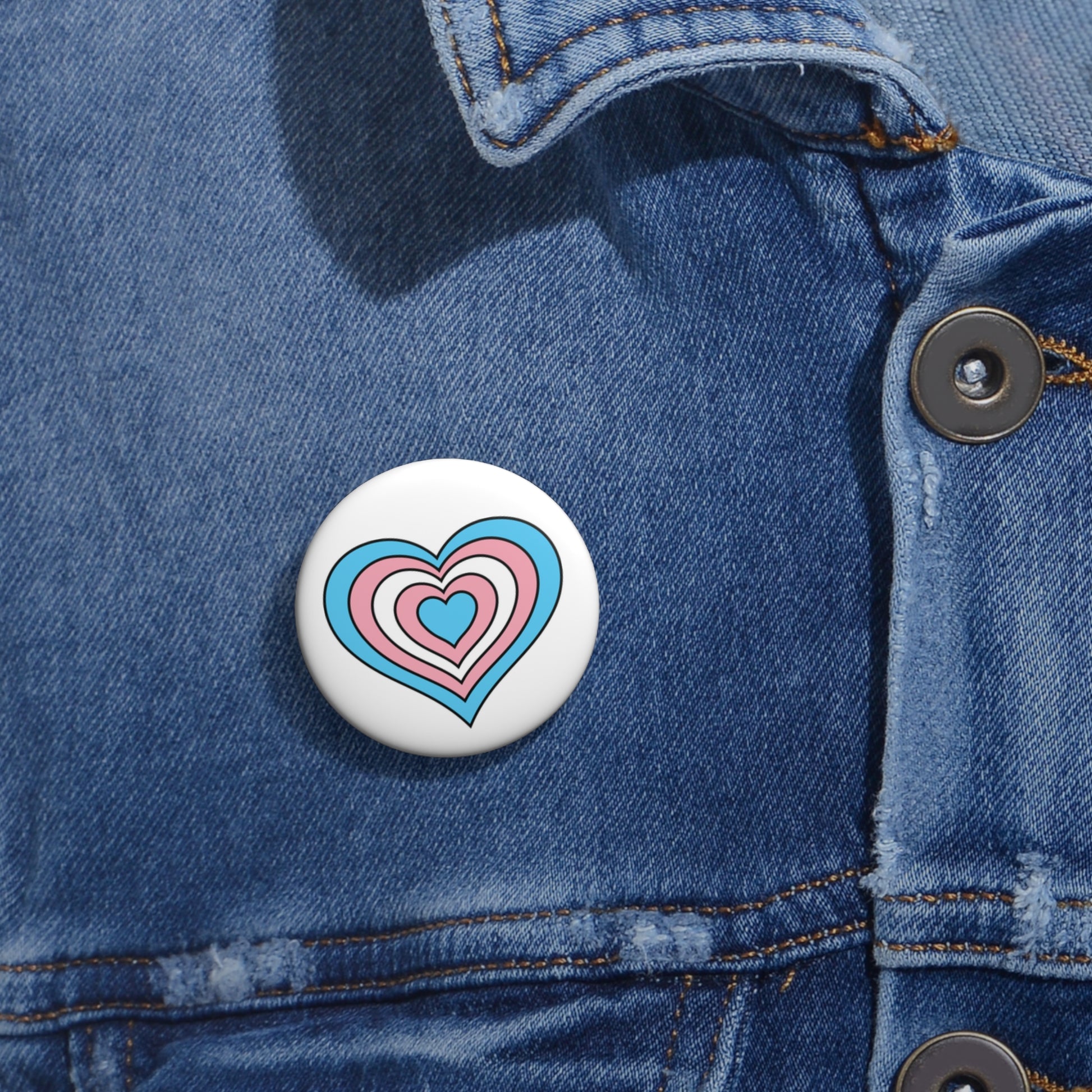 Close-up of a white circular pride button featuring a nested heart design inspired by the Transgender pride flag in blue, pink and white, attached to a blue denim jacket at the right chest with the button clearly visible.