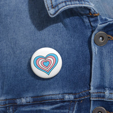 Close-up of a white circular pride button featuring a nested heart design inspired by the Transgender pride flag in blue, pink and white, attached to a blue denim jacket at the right chest with the button clearly visible.