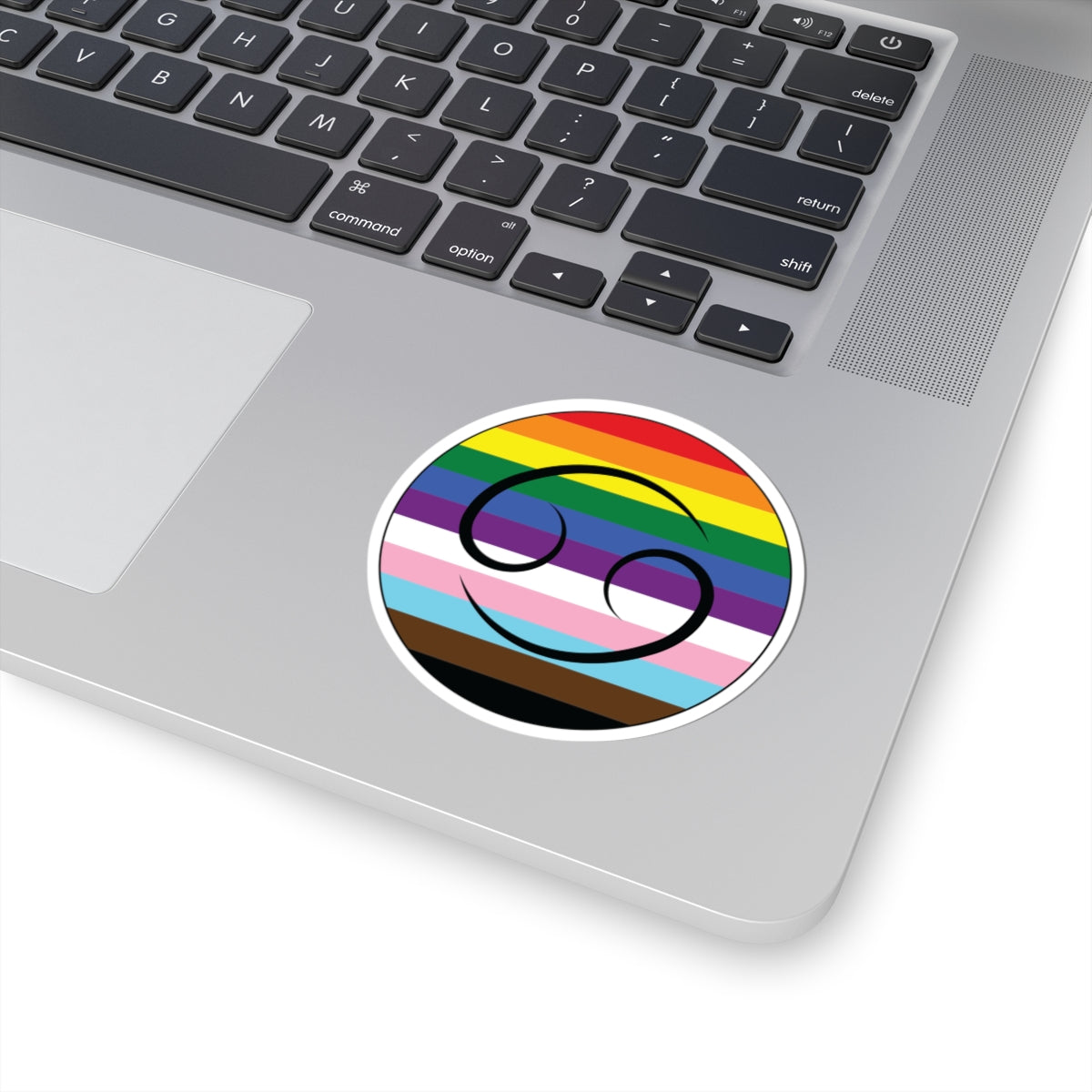 Close-up of the bottom right corner of a silver laptop featuring a 3 inch by 3 inch circular zodiac sticker with a centered Cancer glyph on a progress pride flag style background applied to the surface.