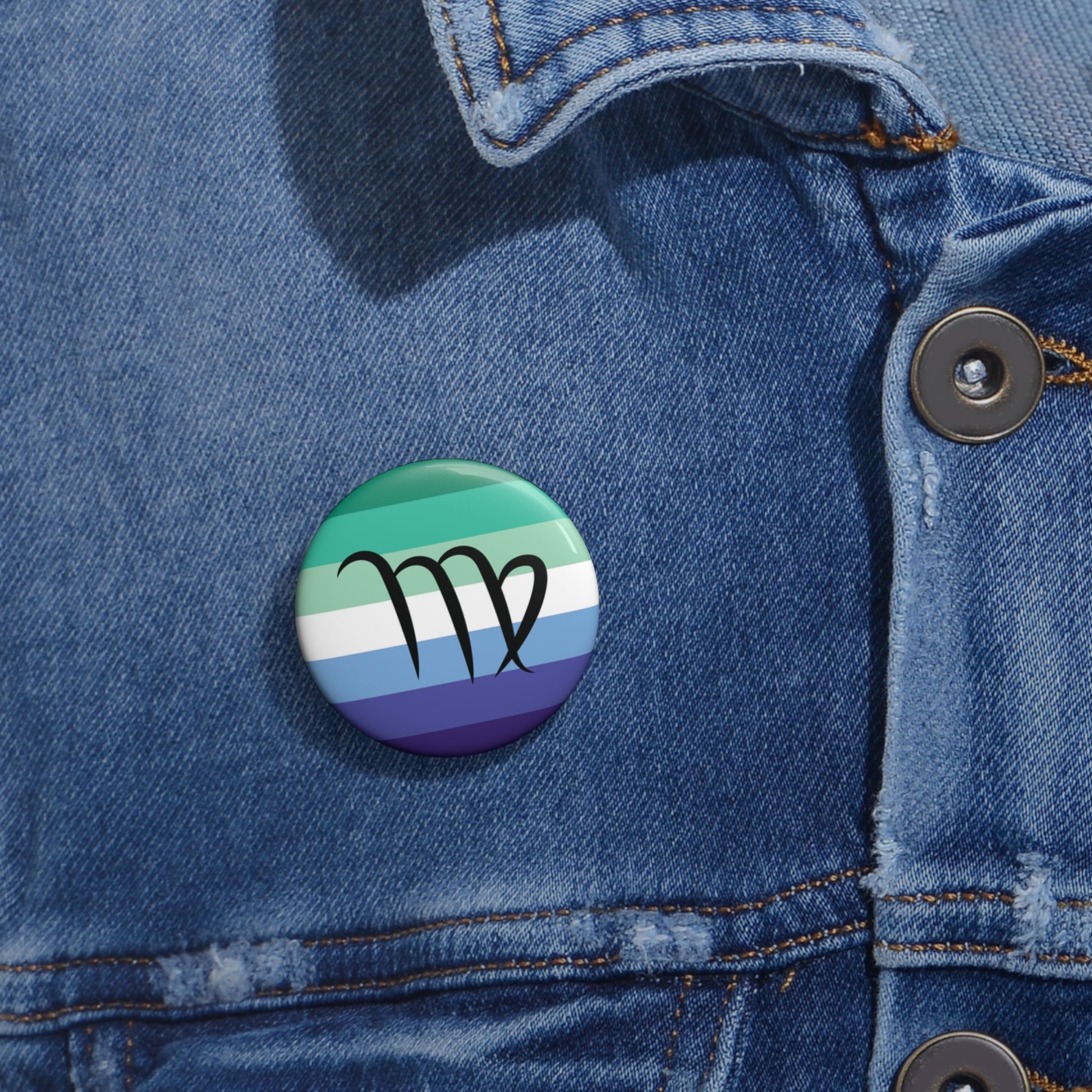 Close-up of a white circular pride button featuring the Virgo zodiac glyph centered over a gay men (MLM) pride flag background in greens/teals, white, and blues, attached to a blue denim jacket at the right chest with the button clearly visible.