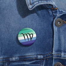 Close-up of a white circular pride button featuring the Virgo zodiac glyph centered over a gay men (MLM) pride flag background in greens/teals, white, and blues, attached to a blue denim jacket at the right chest with the button clearly visible.