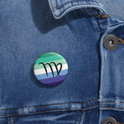 Close-up of a white circular pride button featuring the Virgo zodiac glyph centered over a gay men (MLM) pride flag background in greens/teals, white, and blues, attached to a blue denim jacket at the right chest with the button clearly visible.