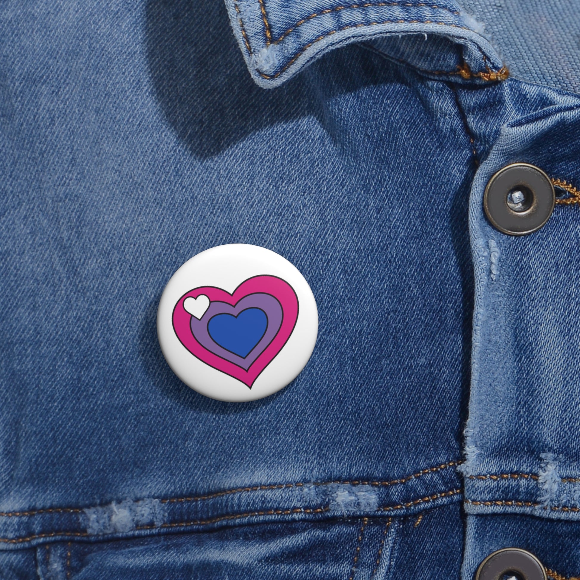Close-up of a white circular pride button featuring a nested heart design inspired by the biromantic pride flag in pink, lavender and blue with a white heart on the left side, attached to a blue denim jacket at the right chest with the button clearly visible.