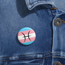 Close-up of a white circular pride button featuring the Pisces zodiac glyph centered over a transgender pride flag background in light blue, pink, and white, attached to a blue denim jacket at the right chest with the button clearly visible.