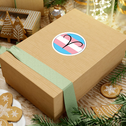 Gift box with a green ribbon on a festive background with cookies and Christmas decorations. Aires Transgender pride sticker centered on the box.