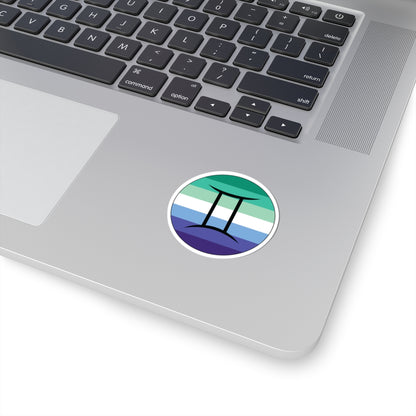 Close-up of the bottom right corner of a silver laptop featuring a 2 inch by 2 inch circular zodiac sticker with a centered Gemini glyph on a Gay Men pride flag style background applied to the surface.