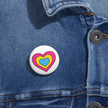 Close-up of a white circular pride button featuring a nested heart design inspired by the Pansexual pride flag in pink, yellow and blue, attached to a blue denim jacket at the right chest with the button clearly visible.