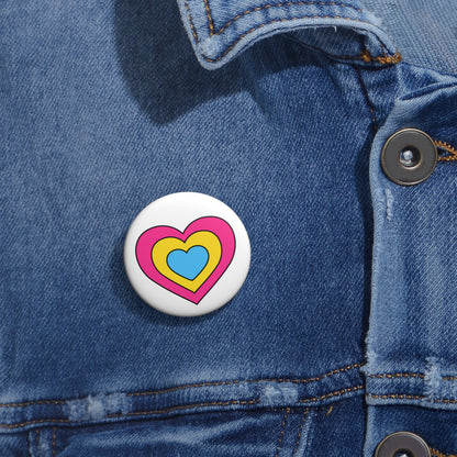 Close-up of a white circular pride button featuring a nested heart design inspired by the Pansexual pride flag in pink, yellow and blue, attached to a blue denim jacket at the right chest with the button clearly visible.