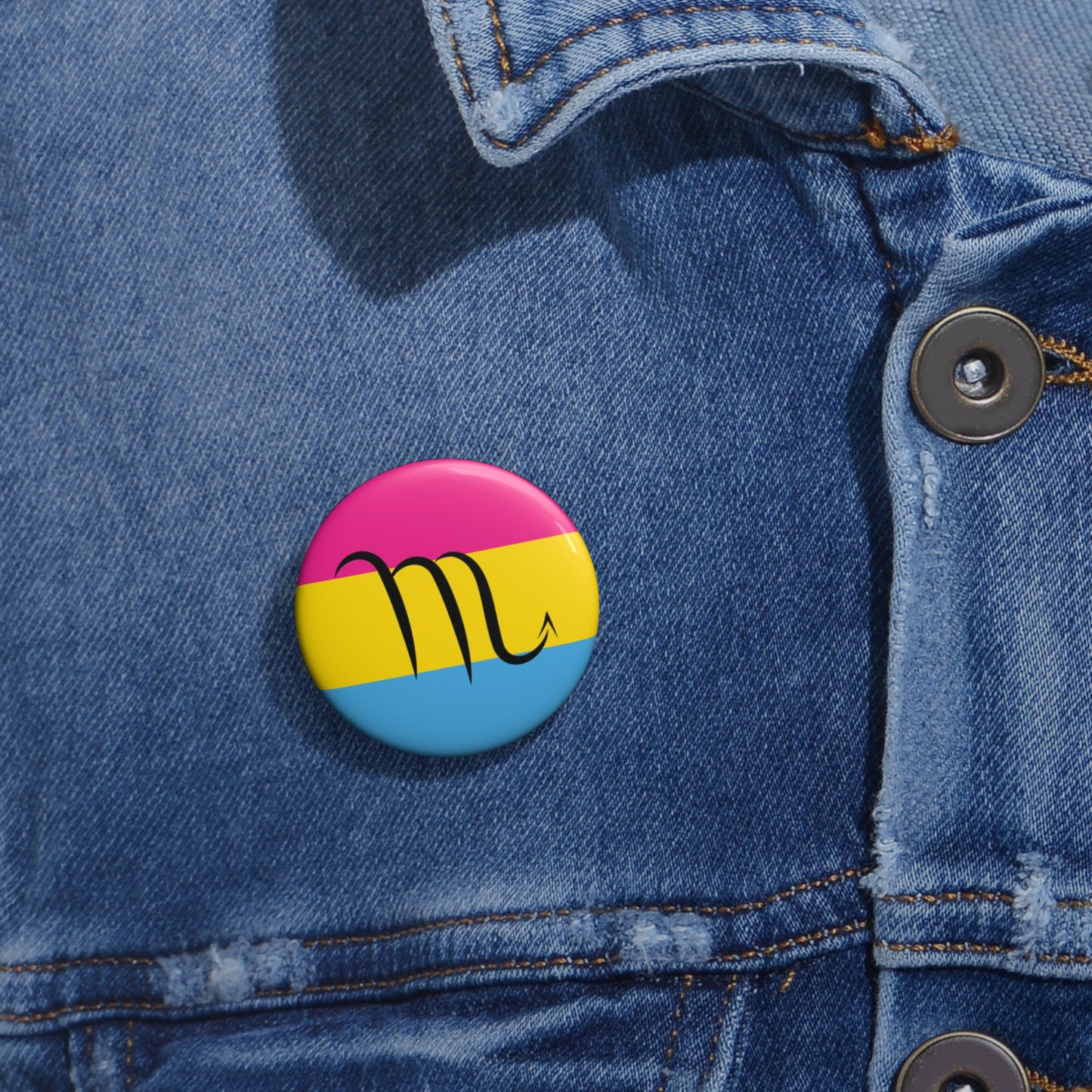 Close-up of a white circular pride button featuring the Scorpio zodiac glyph centered over a Pansexual pride flag background in pink, yellow, and blue, attached to a blue denim jacket at the right chest with the button clearly visible.