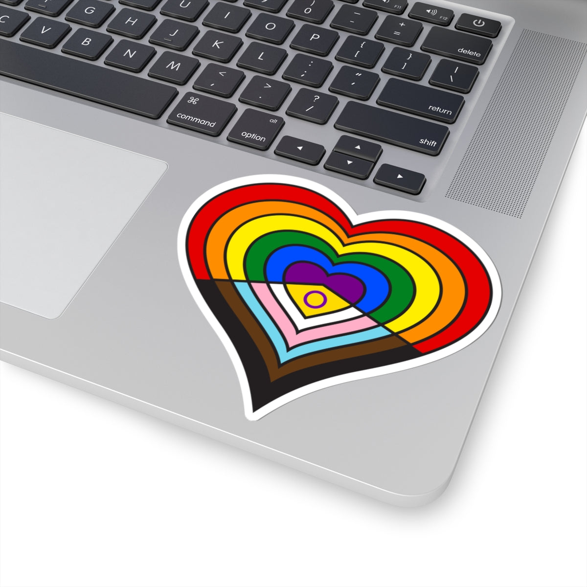 Close-up of the bottom right corner of a silver laptop featuring a 4 inch by 4 inch pride flag heart sticker in an Intersex-Inclusive Progress pride flag style applied to the surface.