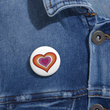 Close-up of a white circular pride button featuring a nested heart design inspired by the Lesbian pride flag in oranges, white and pinks, attached to a blue denim jacket at the right chest with the button clearly visible.