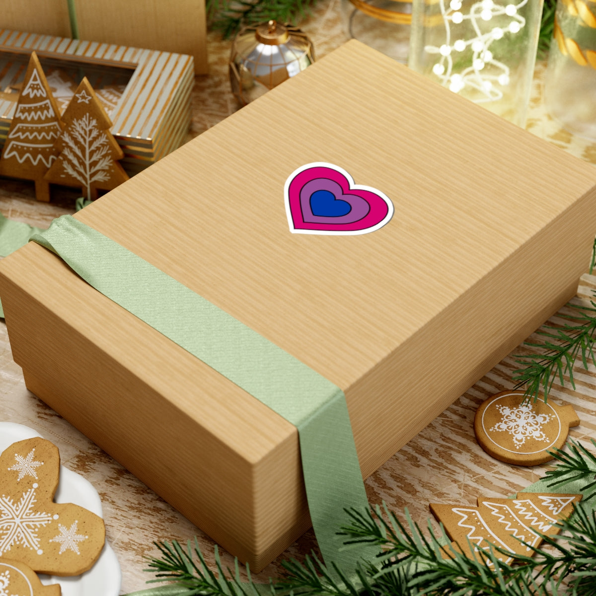 Gift box with a green ribbon on a festive background with cookies and Christmas decorations. Heart-shaped Bisexual pride sticker centered on the box.