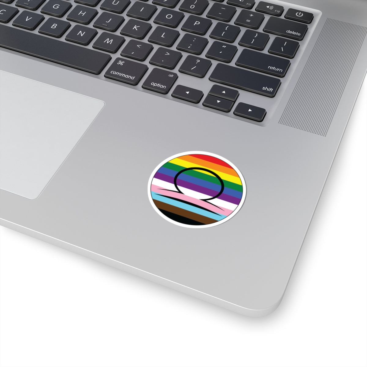 Close-up of the bottom right corner of a silver laptop featuring a 2 inch by 2 inch circular zodiac sticker with a centered Libra glyph on a progress pride flag style background applied to the surface.