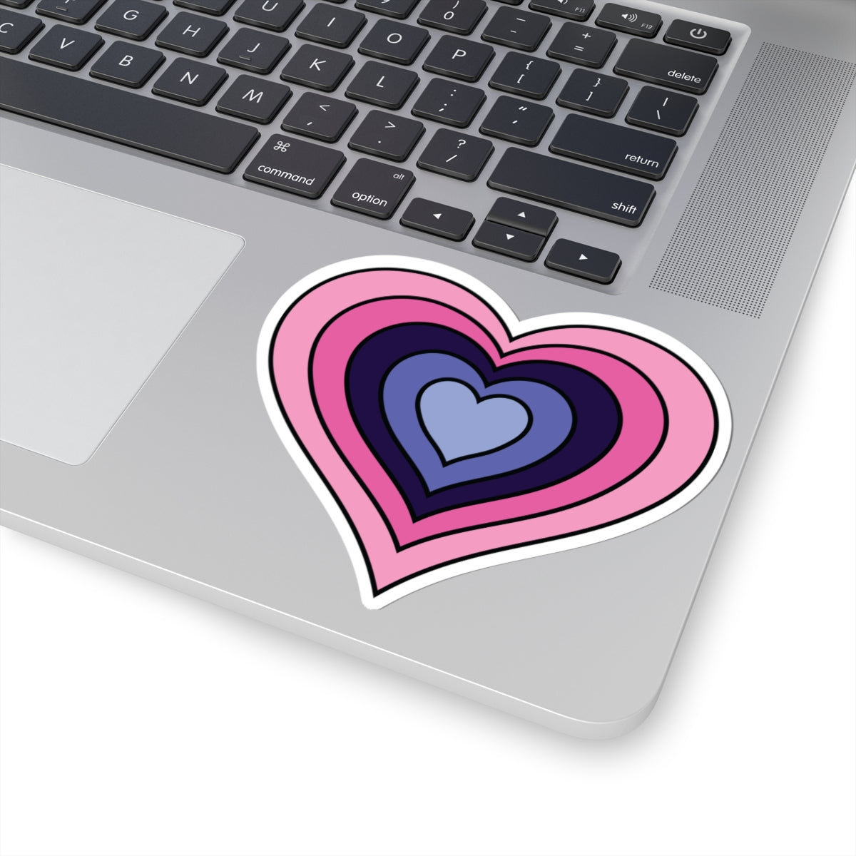 Close-up of the bottom right corner of a silver laptop featuring a 4 inch by 4 inch pride flag heart sticker in an Omnigender flag style applied to the surface.