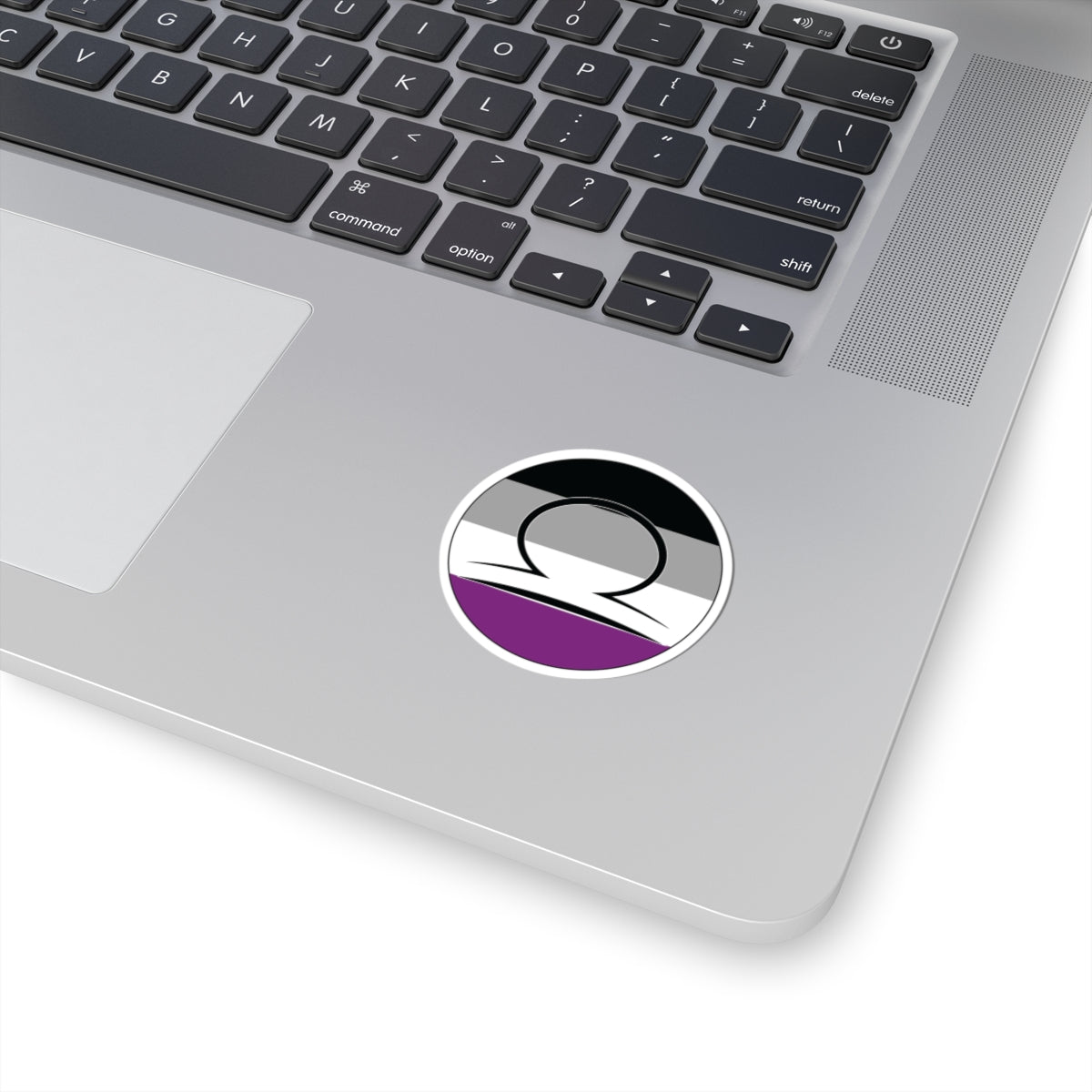Close-up of the bottom right corner of a silver laptop featuring a 2 inch by 2 inch circular zodiac sticker with a centered Libra glyph on an Asexual pride flag style background applied to the surface.