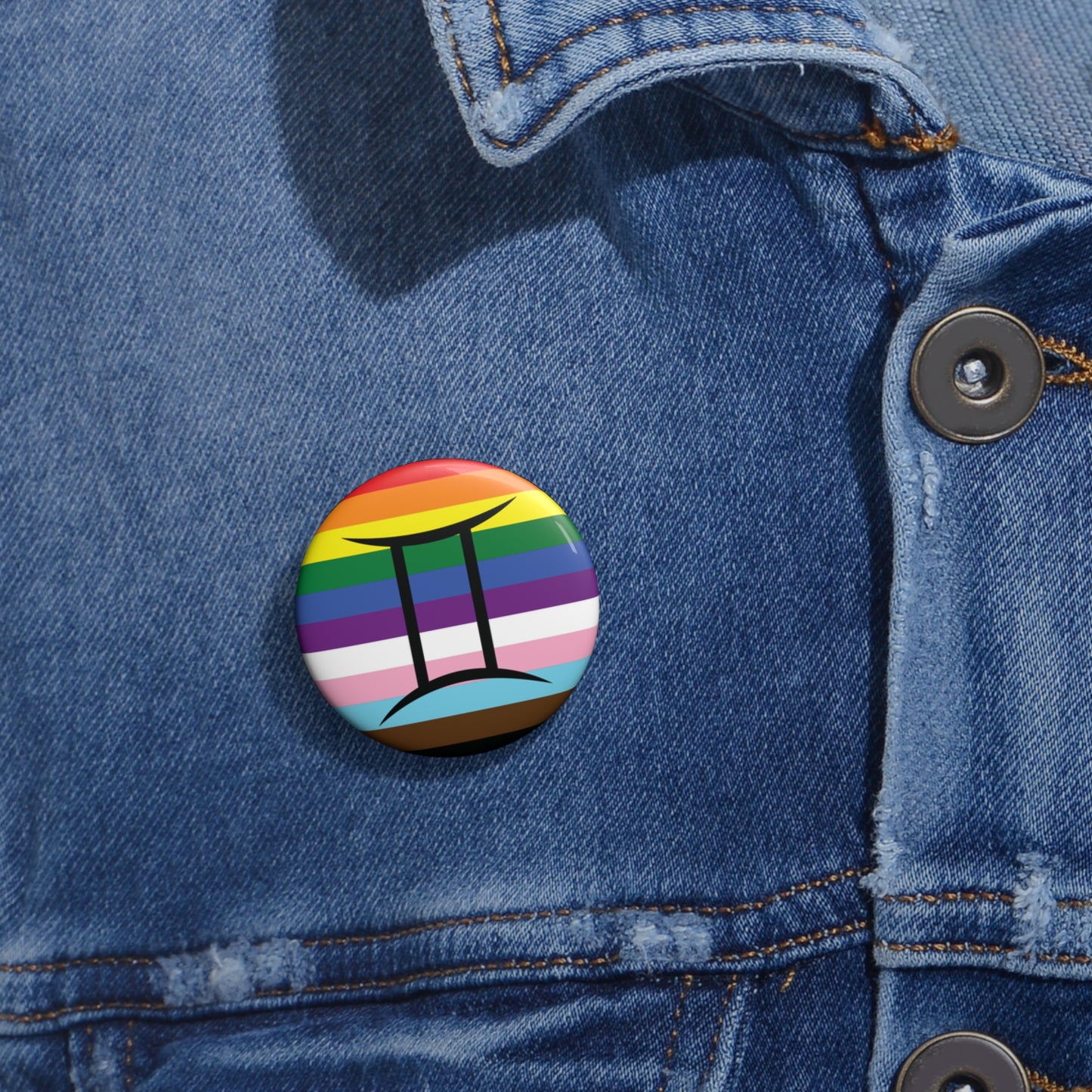 Close-up of a white circular pride button featuring the Gemini zodiac glyph centered over a Progress Pride flag background in rainbow stripes with black, brown, light blue, pink, and white elements, attached to a blue denim jacket at the right chest with the button clearly visible.