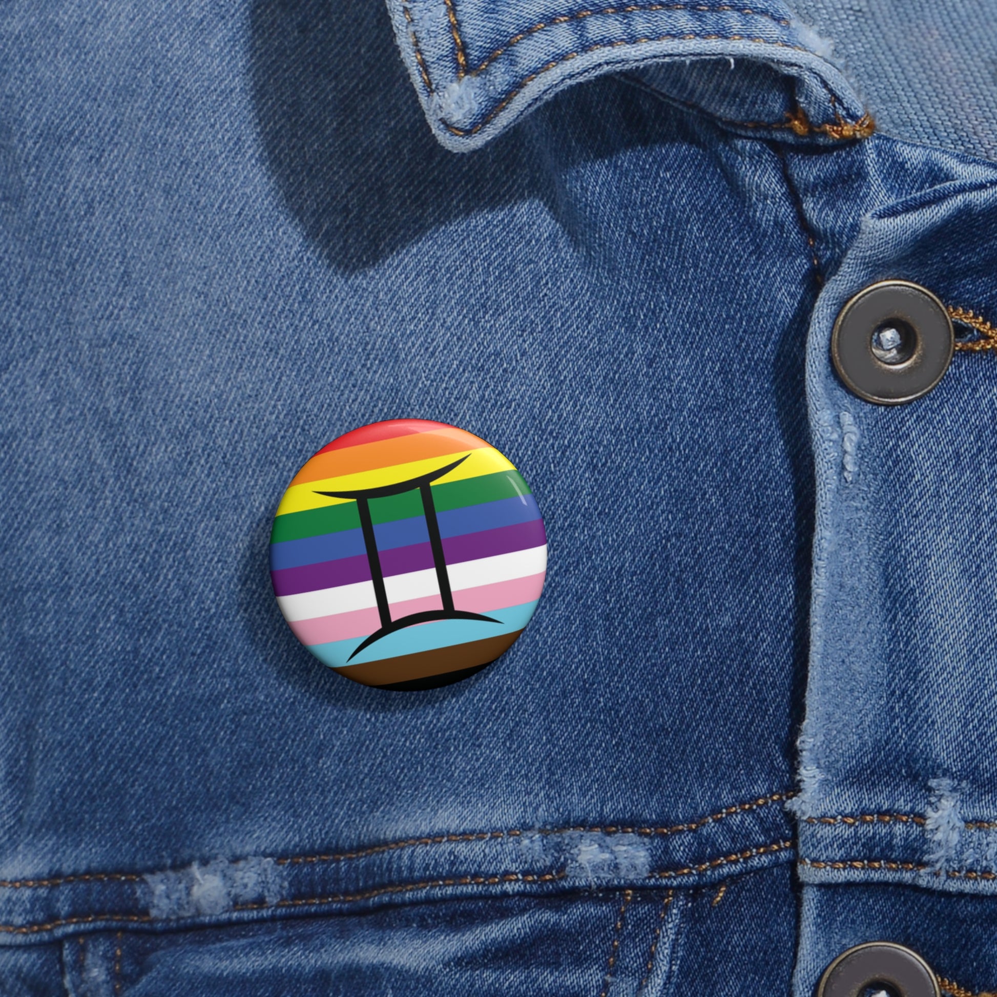 Close-up of a white circular pride button featuring the Gemini zodiac glyph centered over a Progress Pride flag background in rainbow stripes with black, brown, light blue, pink, and white elements, attached to a blue denim jacket at the right chest with the button clearly visible.