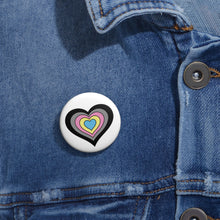 Close-up of a white circular pride button featuring a nested heart design inspired by the Polygender pride flag in black, gray, pink, yellow and blue, attached to a blue denim jacket at the right chest with the button clearly visible.