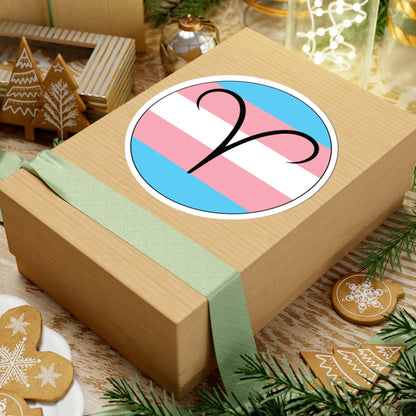 Gift box with a green ribbon on a festive background with cookies and Christmas decorations. Aires Transgender pride sticker centered on the box.