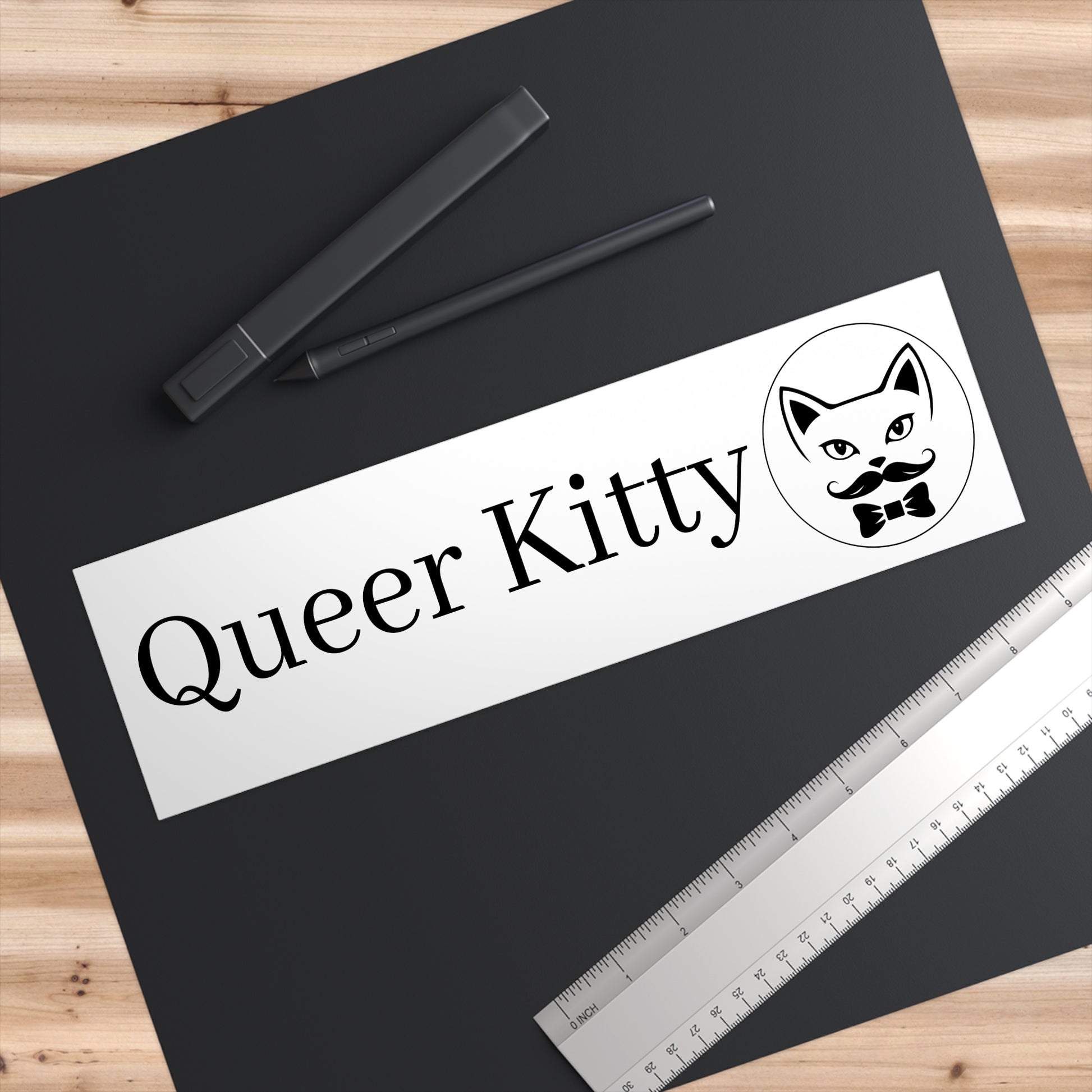 Black paper with 'Queer Kitty' text and logo on a wooden surface with pens and a ruler.
