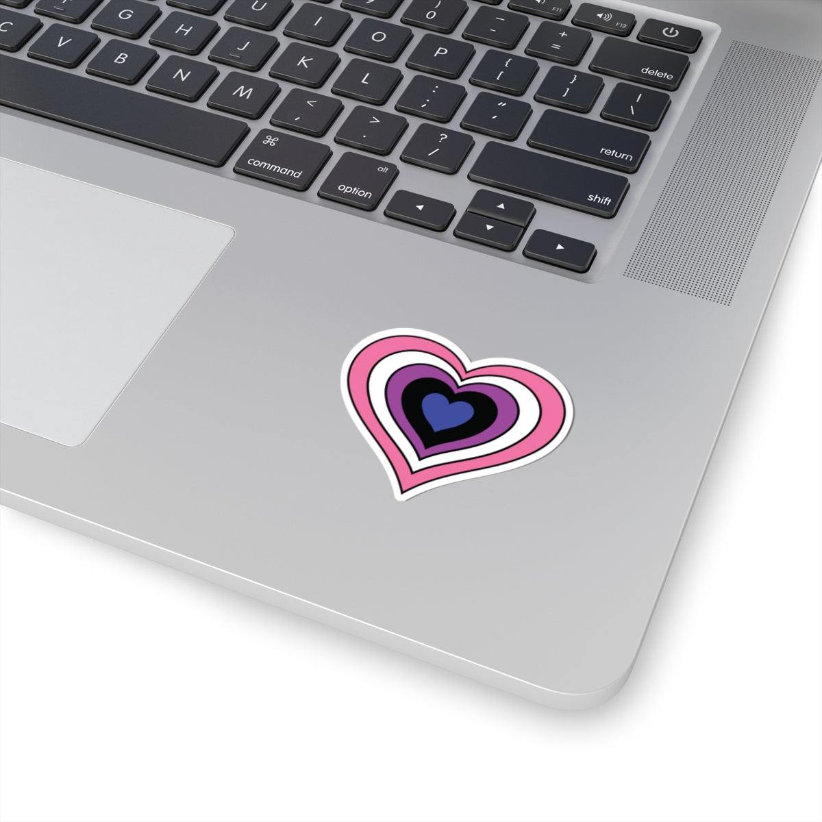Close-up of the bottom right corner of a silver laptop featuring a 2 inch by 2 inch pride flag heart sticker in a Genderfluid flag style applied to the surface.