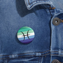 Close-up of a white circular pride button featuring the Pisces zodiac glyph centered over a gay men (MLM) pride flag background in greens/teals, white, and blues, attached to a blue denim jacket at the right chest with the button clearly visible.