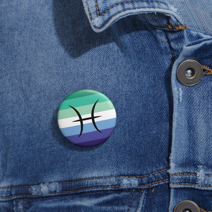 Close-up of a white circular pride button featuring the Pisces zodiac glyph centered over a gay men (MLM) pride flag background in greens/teals, white, and blues, attached to a blue denim jacket at the right chest with the button clearly visible.