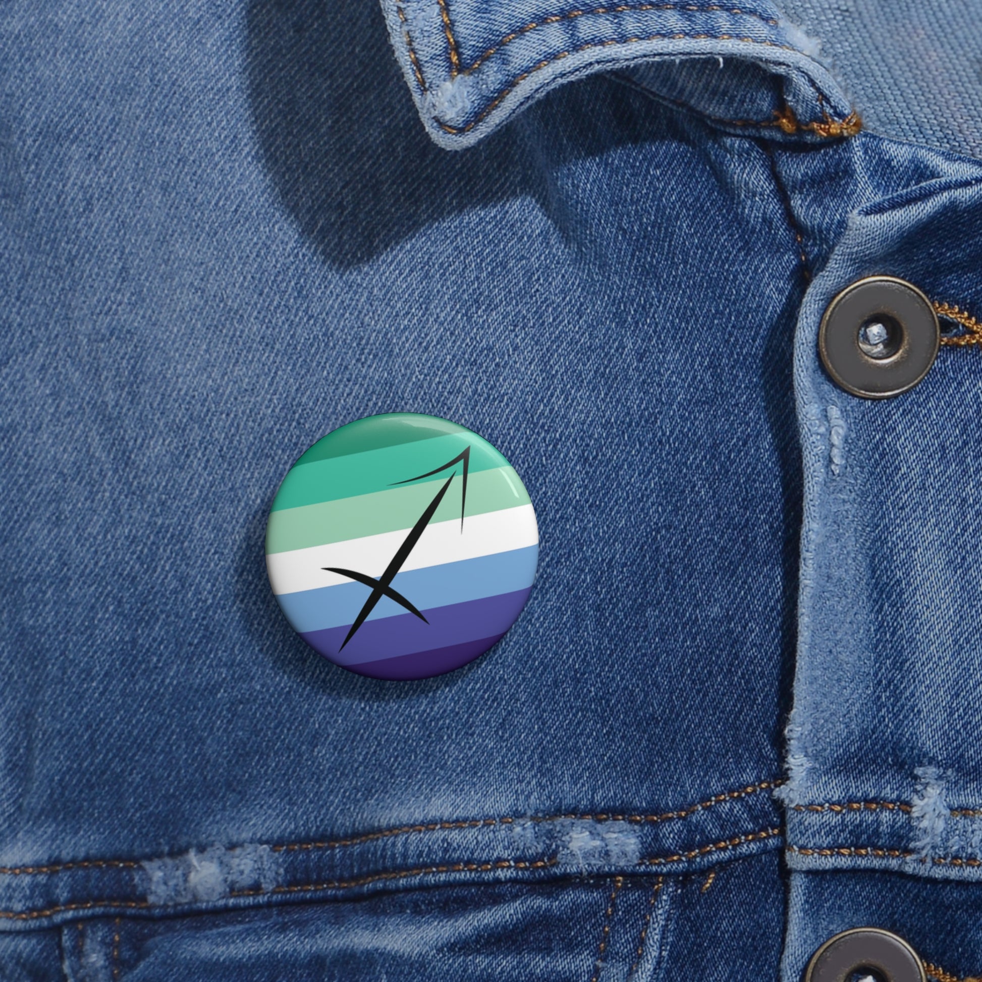 Close-up of a white circular pride button featuring the Sagittarius zodiac glyph centered over a gay men (MLM) pride flag background in greens/teals, white, and blues, attached to a blue denim jacket at the right chest with the button clearly visible.